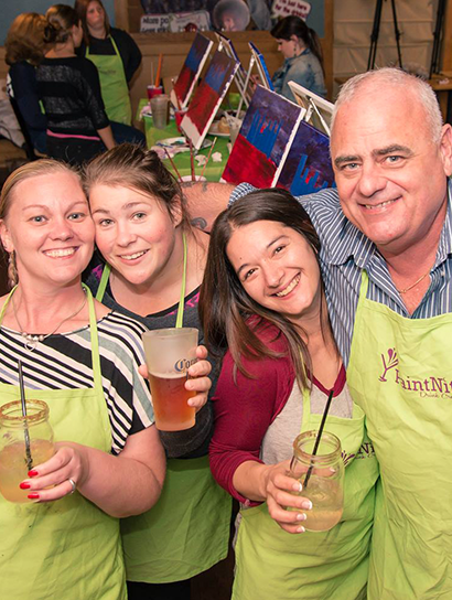 Mom, Dad and daughters smiling in Paint Nite aprons with drinks